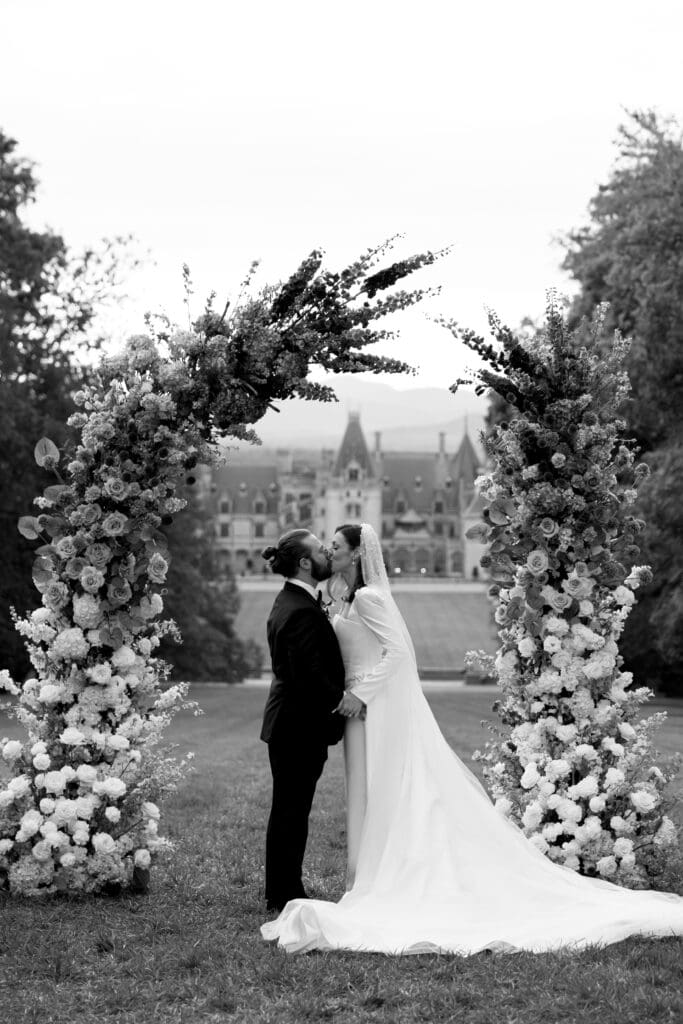 Editorial wedding portrait of a couple kissing under a floral arch on the Diana Lawn with the Biltmore Estate behind them, photographed by a Biltmore wedding photographer.