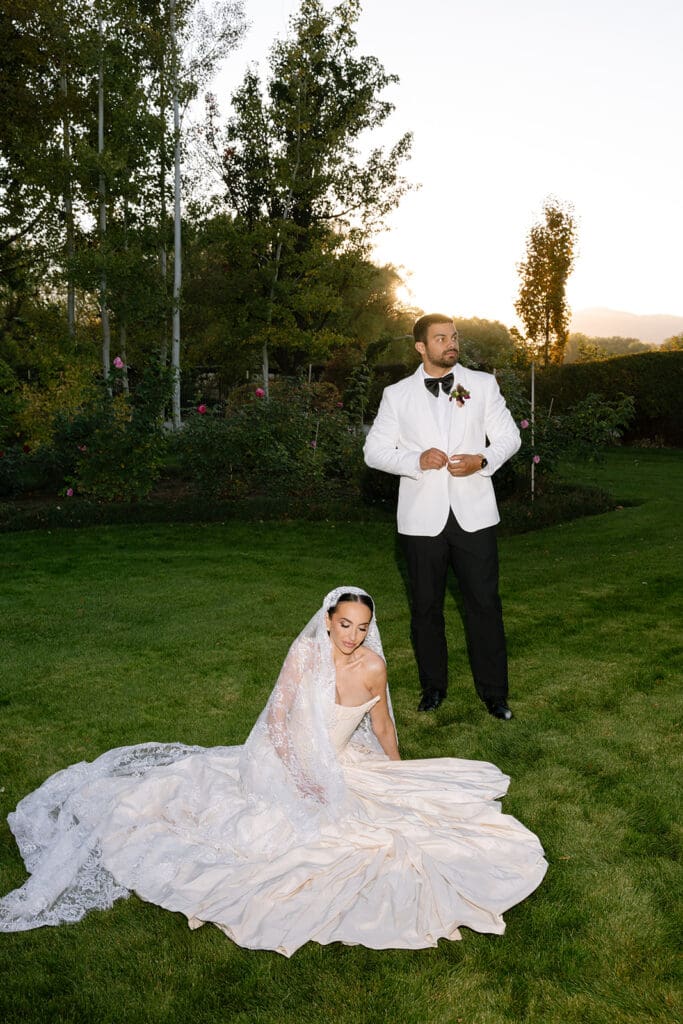 Bride seated in gown on lawn with groom standing behind at Oceana’s Gardens.