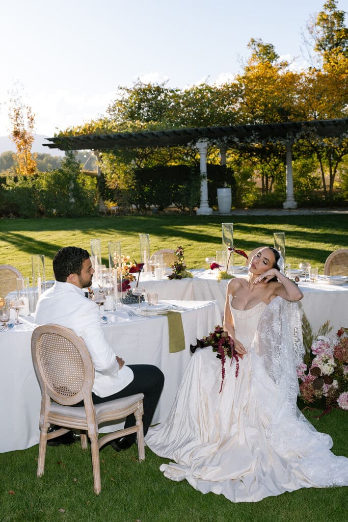 Bride leaning back in chair beside groom at outdoor reception at Oceana’s Gardens.