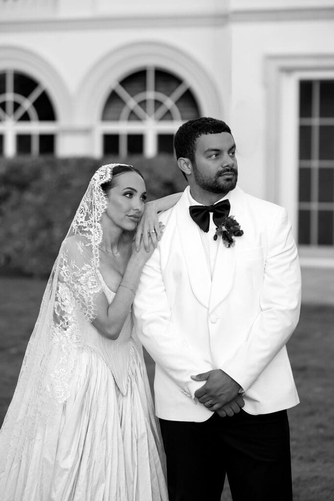 Black and white portrait of bride and groom standing near estate architecture at Oceana’s Gardens.