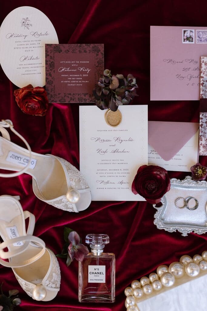 Editorial wedding flat lay with invitations, designer jimmy choo heels, perfume, and deep red florals styled for Santa Barbara wedding.