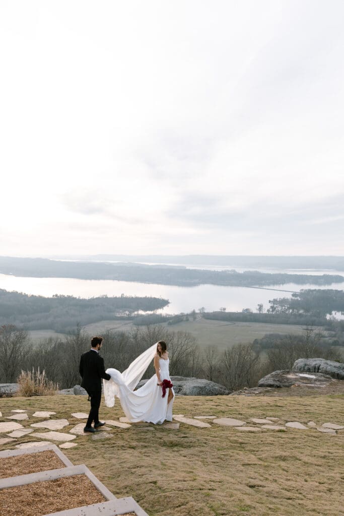 Bride holding a deep red bouquet as the groom trails behind holding her veil at Stone Haven wedding venue in Alabama overlooking the valley and river.