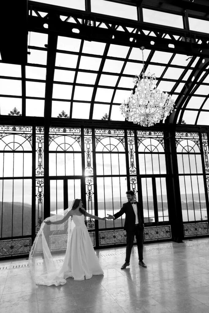Black and white editorial wedding portrait of bride and groom reaching for each other beneath chandeliers inside the Stone Haven glass conservatory in Alabama.