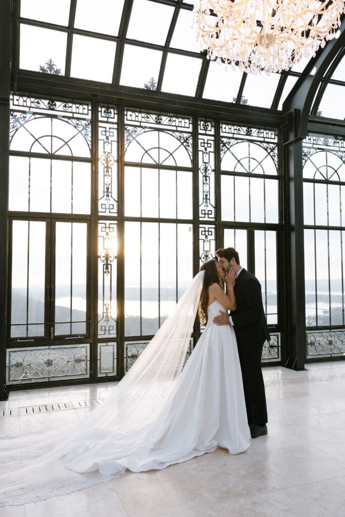 Bride and groom sharing an intimate kiss beneath crystal chandeliers inside the glass conservatory at Stone Haven wedding venue in Alabama with sunset light pouring through iron windows.