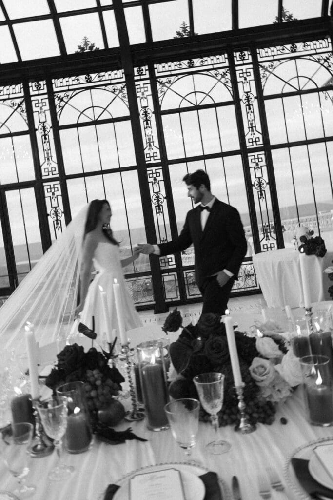 Black and white editorial wedding photo of bride and groom walking hand in hand past candlelit reception tables inside Stone Haven wedding venue in Alabama.
