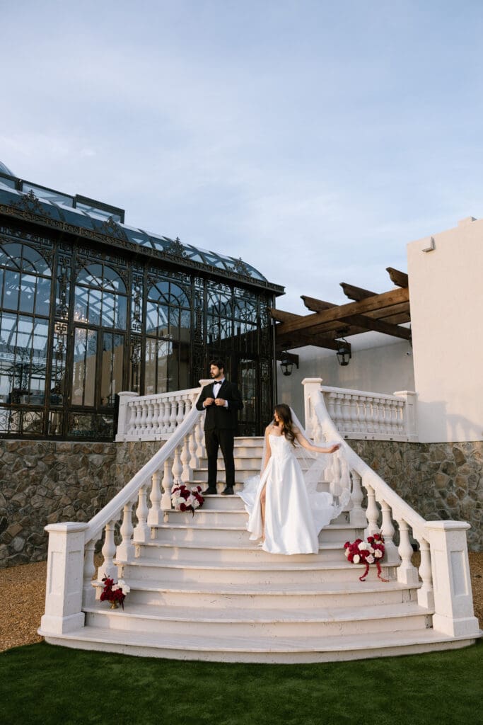 Wide exterior portrait of bride and groom standing on the grand stone staircase outside Stone Haven wedding venue in Alabama with glass conservatory architecture behind them.