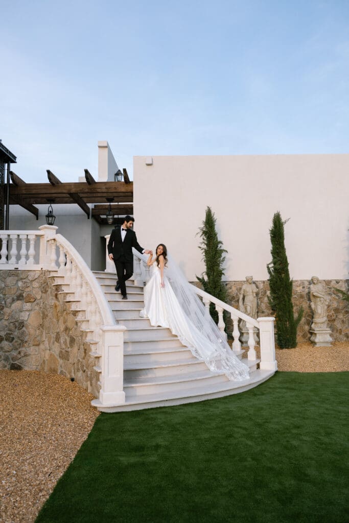 Bride being guided down the staircase by her groom as her cathedral veil trails behind at Stone Haven wedding venue in Alabama during golden hour.