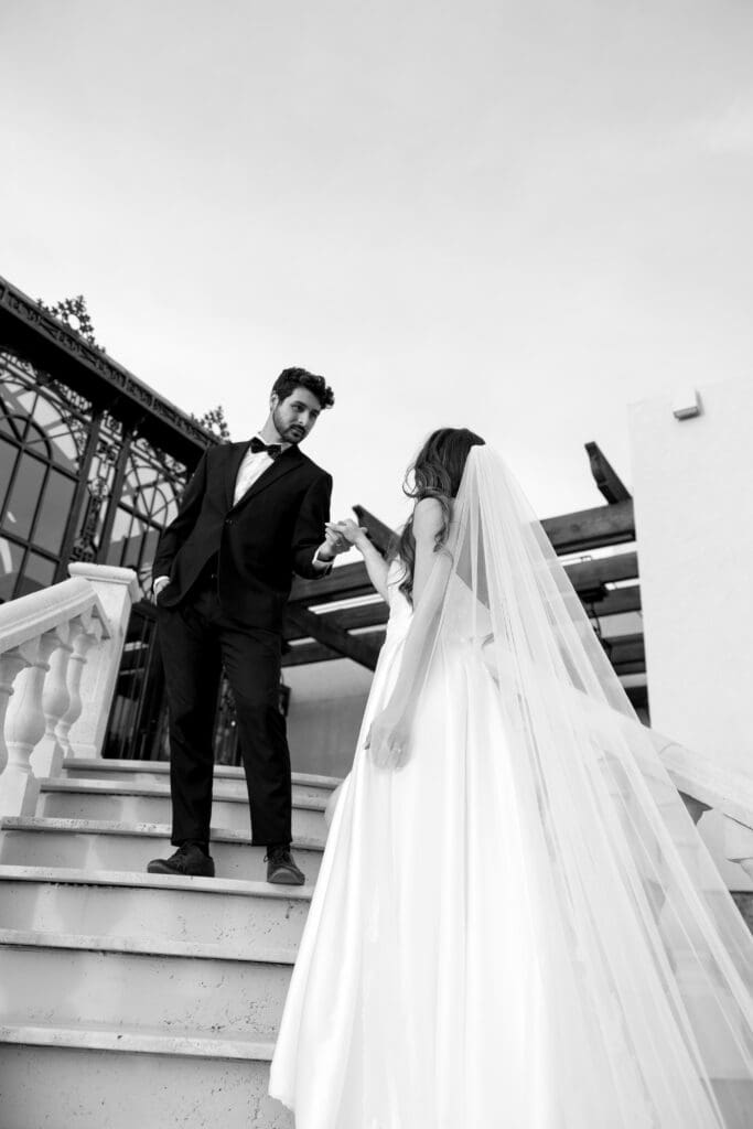 Black and white editorial wedding portrait of groom guiding bride down the grand staircase at Stone Haven wedding venue in Alabama with architectural drama.