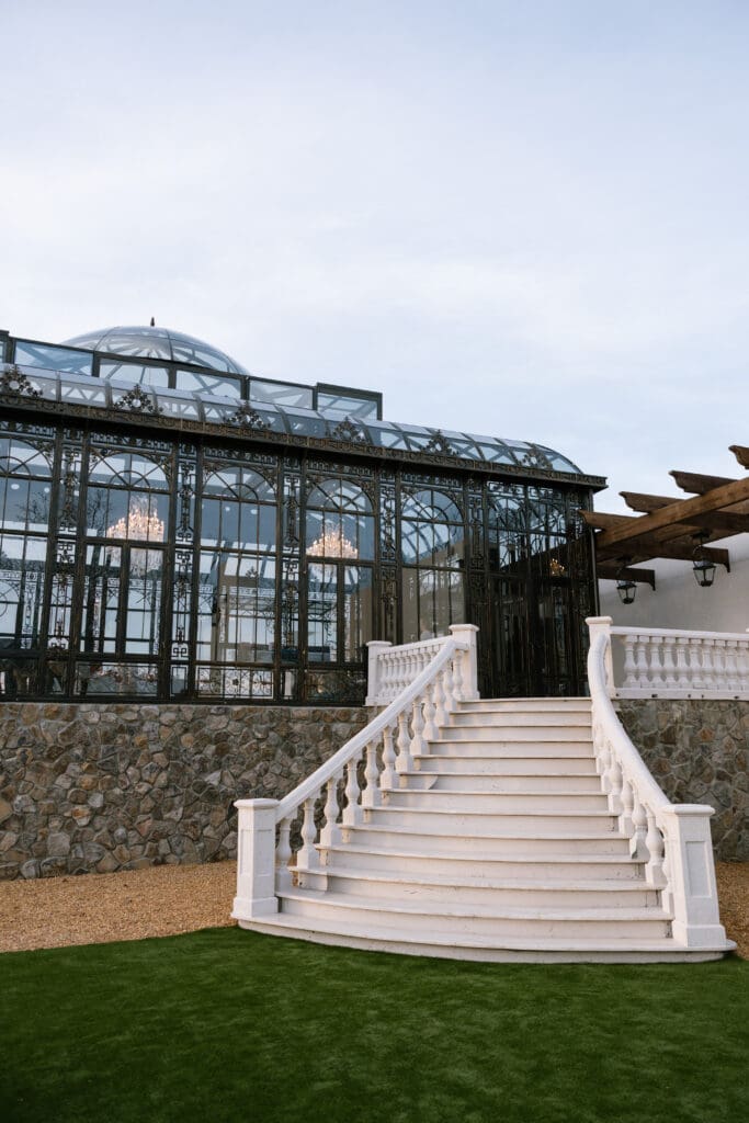 Wide architectural view of the glass conservatory and curved staircase at Stone Haven wedding venue in Alabama showcasing European-inspired ironwork and stone textures.