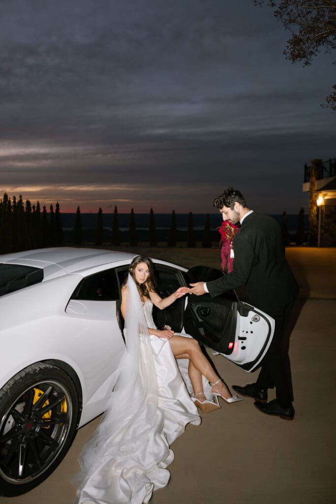 Bride seated at the open door of a white luxury Lamborghini as groom holds her hand at dusk during an editorial wedding moment at Stone Haven wedding venue in Alabama.