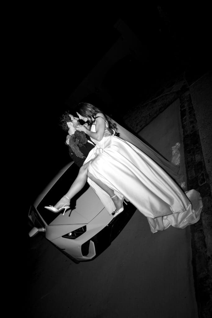Dramatic black and white editorial portrait of groom lifting bride in front of a white luxury Lamborghini at night for a high-fashion wedding moment.