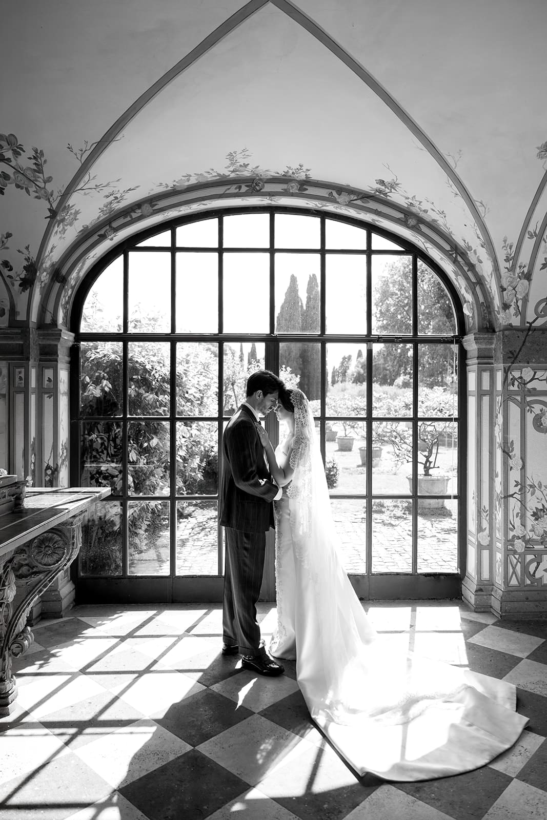 Black and white architectural photograph of bride and groom in classical arched conservatory doorway with geometric floor and botanical backdrop at Rome venue
