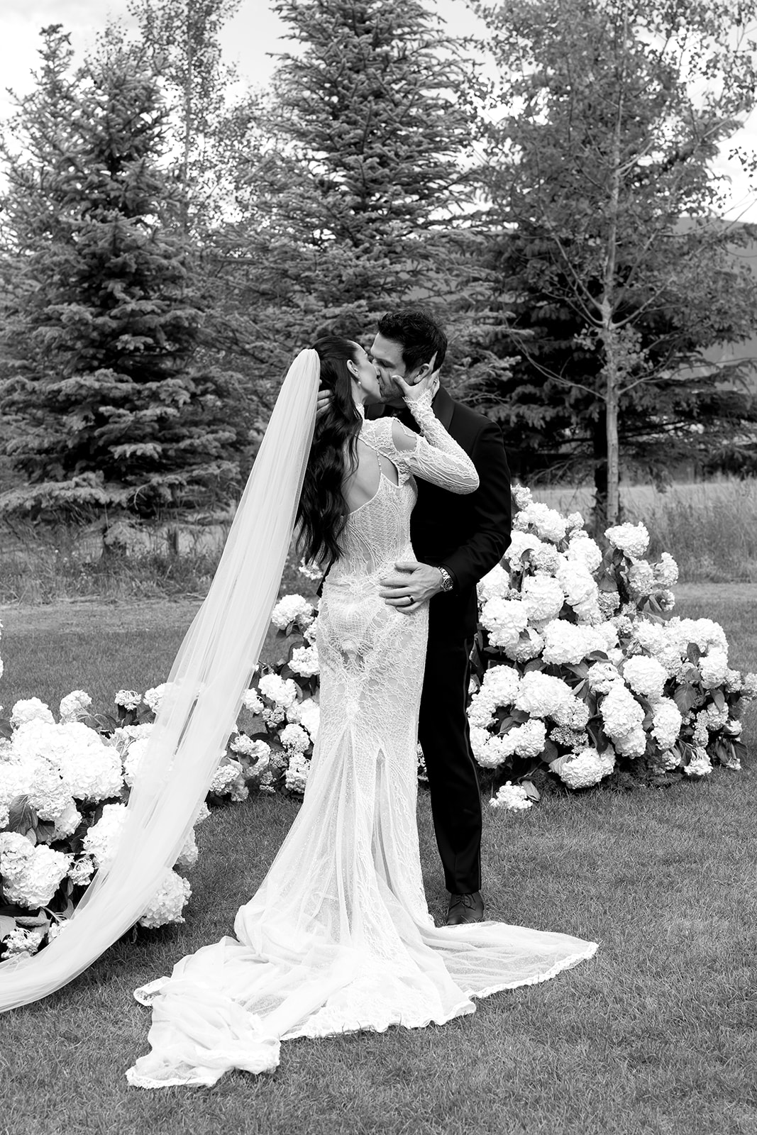 Black and white photo of bride and groom kissing at Montage Big Sky with veil trailing across the lawn and a white hydrangea floral base