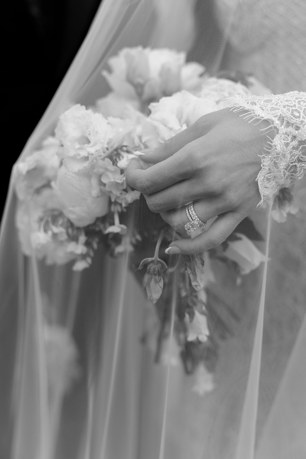 Black and white detail shot of bride's hand holding a bouquet and wedding ring under a cathedral veil