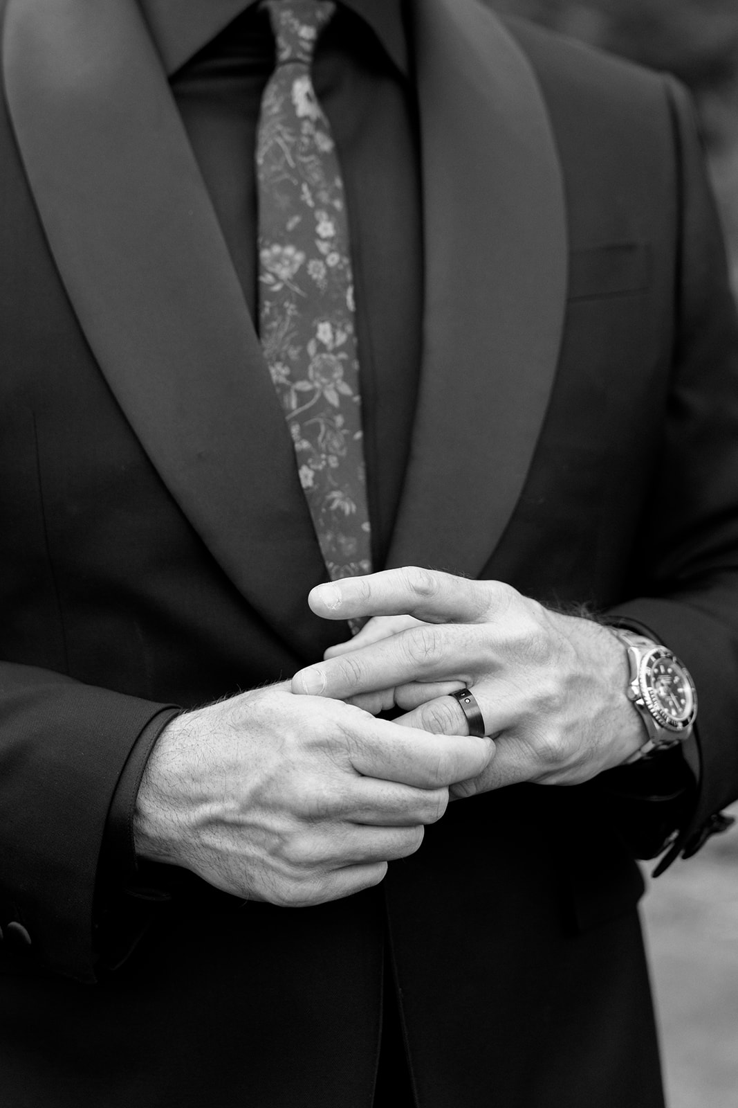Black and white detail of groom's clasped hands showing wedding band and watch with a black tuxedo and patterned tie