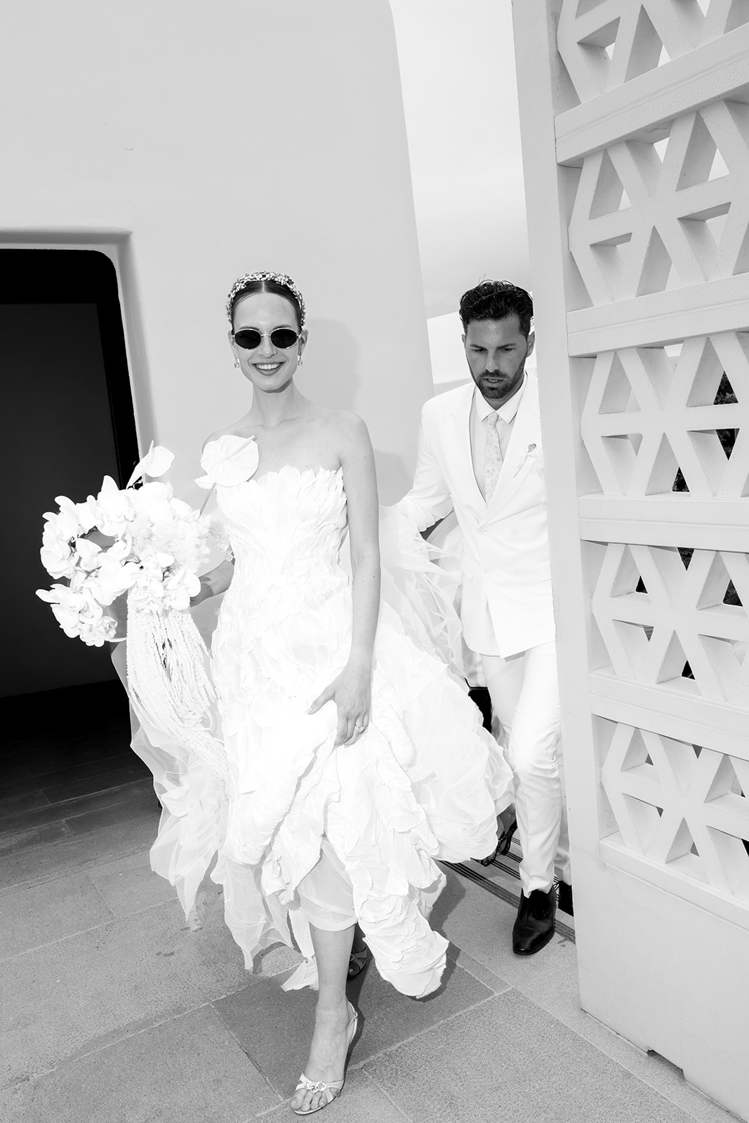 Black and white editorial portrait of a fashionable bride and groom wearing sunglasses beside white architectural details.