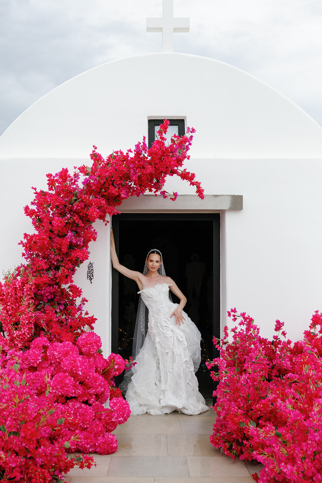 Bride standing beneath a dramatic pink floral installation outside a traditional whitewashed Greek island chapel.