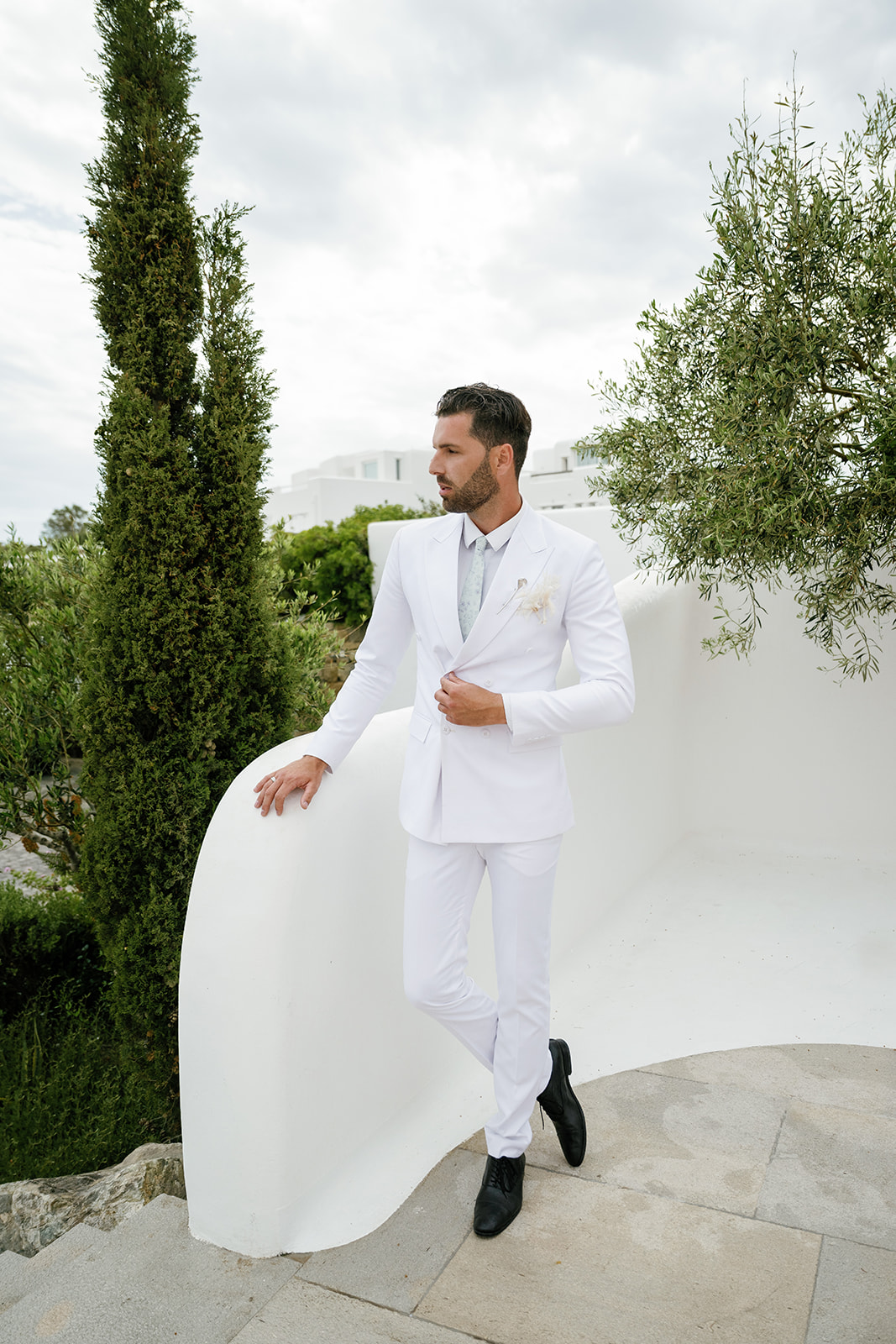 Groom wearing a tailored white suit standing among Mediterranean greenery during a luxury Mykonos wedding.