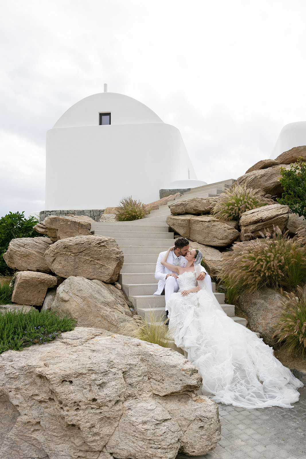 Bride and groom seated among natural stone landscaping with views of the Greek island coastline.
