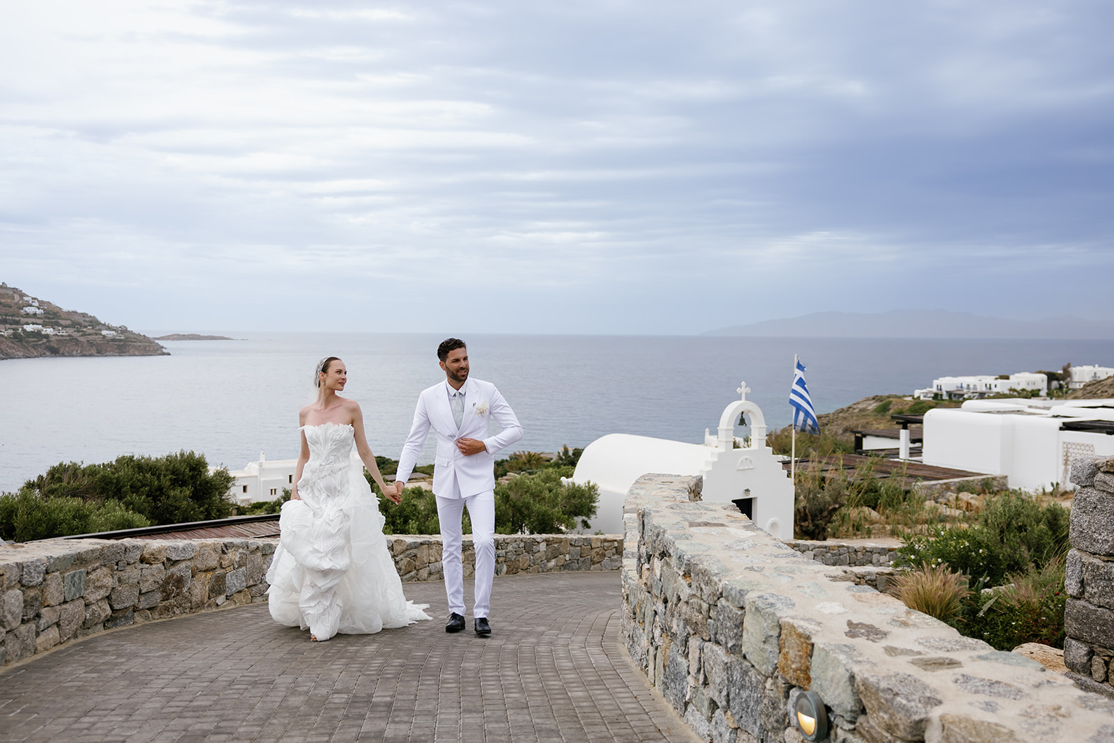 Bride and groom walking together along a whitewashed terrace overlooking the Aegean Sea at a Mykonos destination wedding.