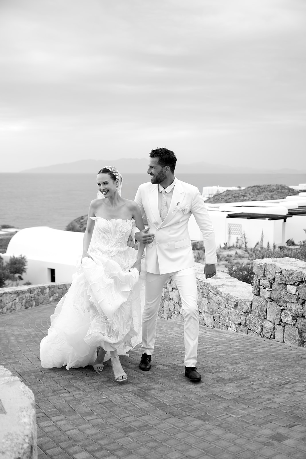 Black and white wedding portrait of a couple embracing with the Greek island coastline in the background.