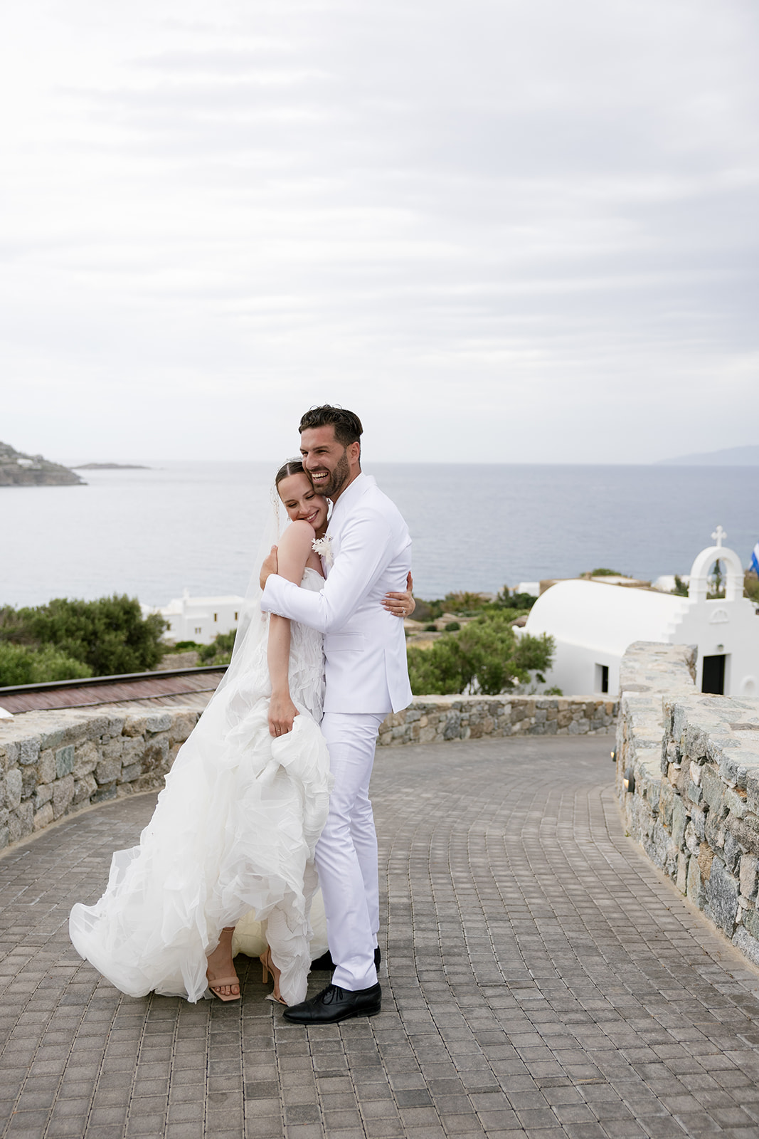 Bride and groom embracing against white Cycladic architecture and coastal views during their luxury Mykonos wedding.