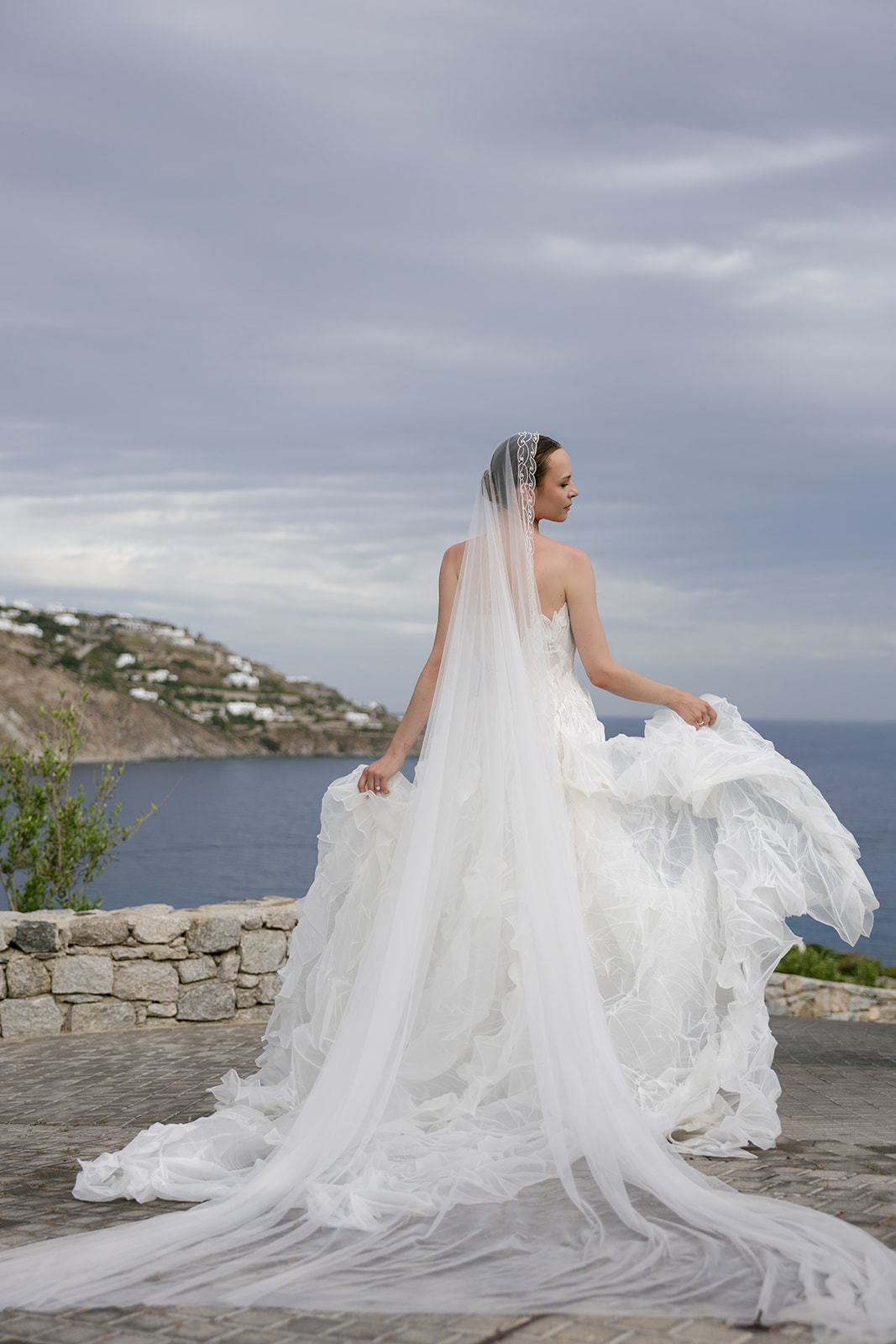 Bride in a flowing white wedding gown overlooking the Aegean Sea during an editorial destination wedding in Mykonos.